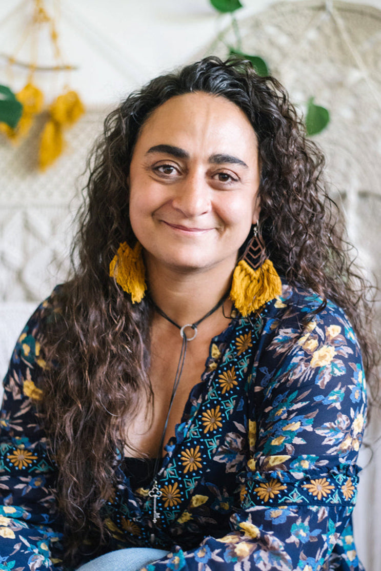 Woman with long curly hair wearing a floral blouse and yellow earrings, sitting in front of a textured wall with plants.