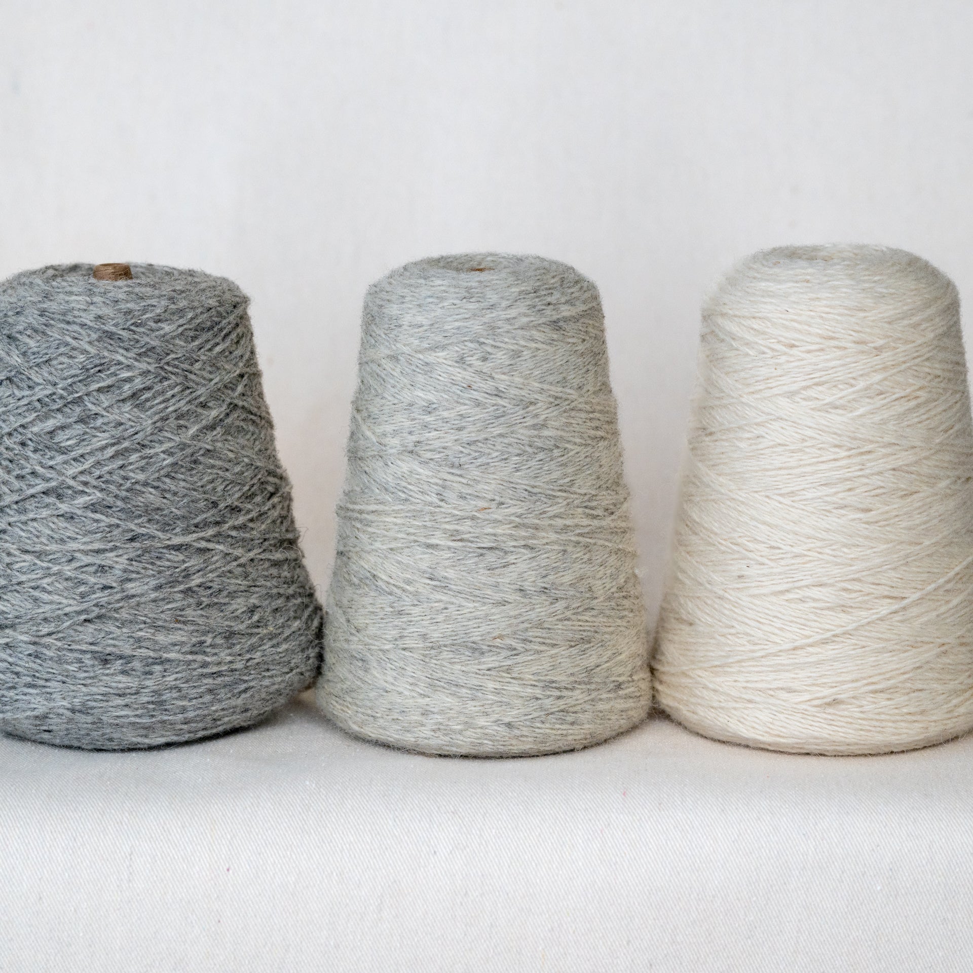Three cones of yarn in gray, white, and beige on a white background